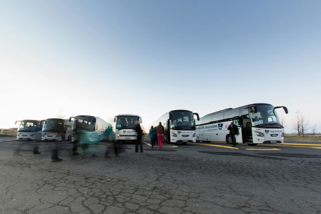 흰색 투어 버스 몇 대가 금이 간 포장 도로에 주차되어 있으며, 사람들은 맑고 연한 푸른 하늘 아래를 거닐고 있습니다.