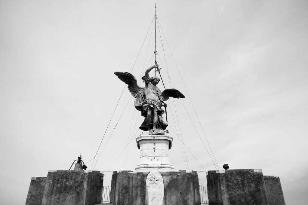 검은색과 흰색의 돌 난간과 케이블로 둘러싸인 받침대에 칼을 들고 있는 날개 달린 형상의 조각상.