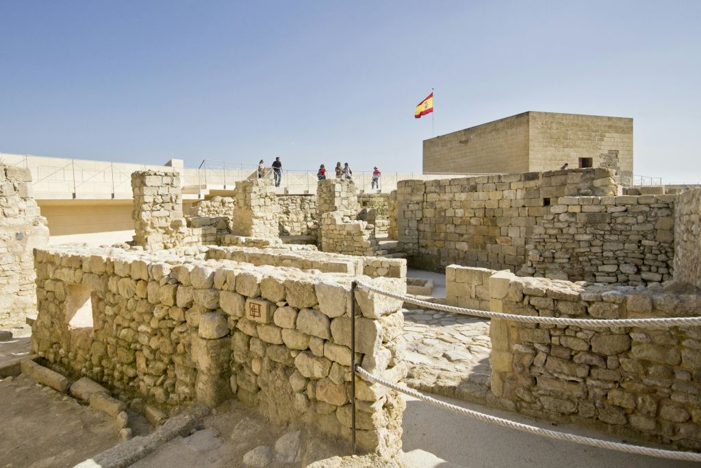 위의 통로를 걷는 사람들의 무리와 함께 고대 석조 유적. 맑은 하늘 아래 배경에 스페인 국기가 보입니다.
