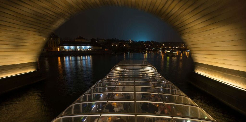 밤에는 강가에 식탁이 놓인 유리 지붕의 보트에 불이 켜진 건물과 반사가 배경으로 보입니다.