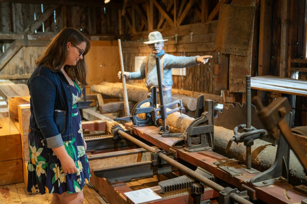 꽃무늬 드레스를 입은 한 여성이 소박한 박물관에서 나무 기계를 관찰하고 있으며, 마네킹이 통나무 커팅을 시연하고 있습니다.