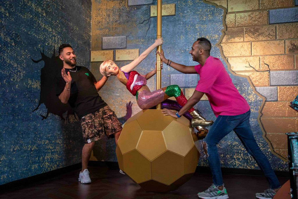 질감이 있는 벽이 있는 테마가 있는 방에서 두 남자가 망가진 공을 든 여성의 왁스 형상과 상호 작용합니다.