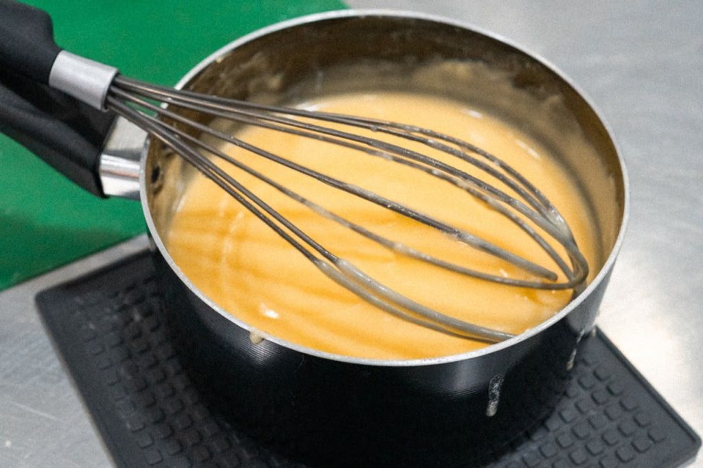 크리미한 노란색 혼합물을 채운 소스팬을 휘저어 주방의 질감 있는 표면에 놓습니다.