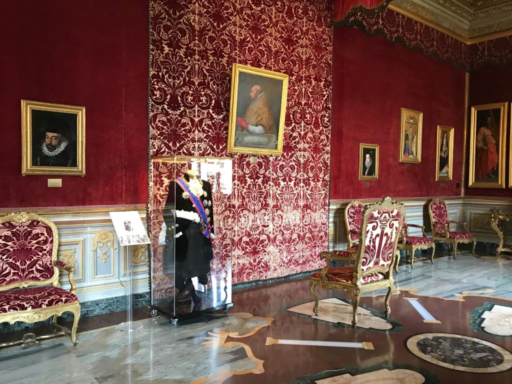 붉은 다마스크 벽, 액자 초상화, 유리로 둘러싸인 유니폼, 금색 디테일이 돋보이는 호화로운 의자가 있는 화려한 객실. 대리석 바닥.
