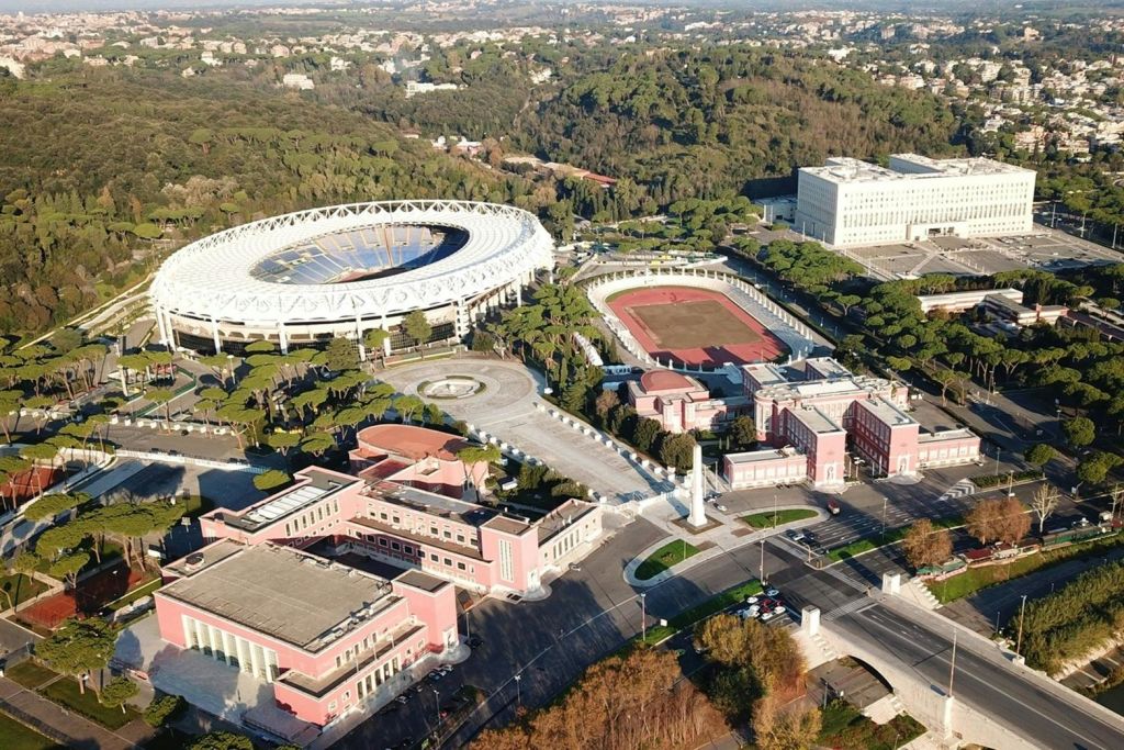 대형 경기장, 트랙 필드, 주변의 여러 건물과 나무가 있는 스포츠 단지의 공중 전망.