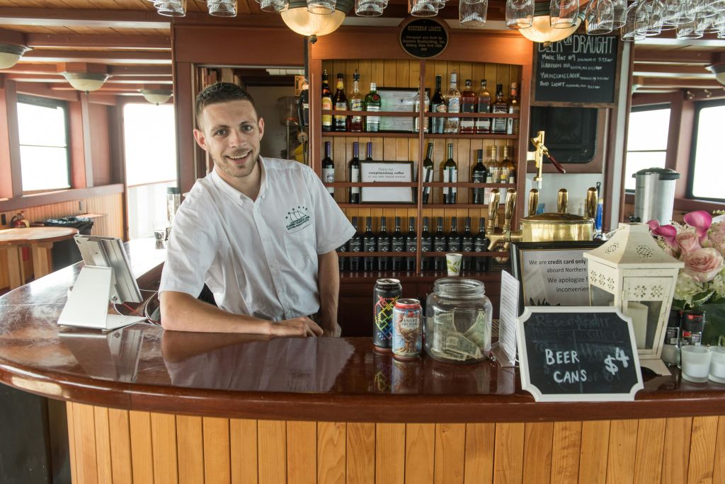 흰색 셔츠를 입은 미소를 짓는 바텐더가 나무 바 뒤에 서 있으며, 카운터에는 술병, 팁 병, 캔이 놓여 있습니다.