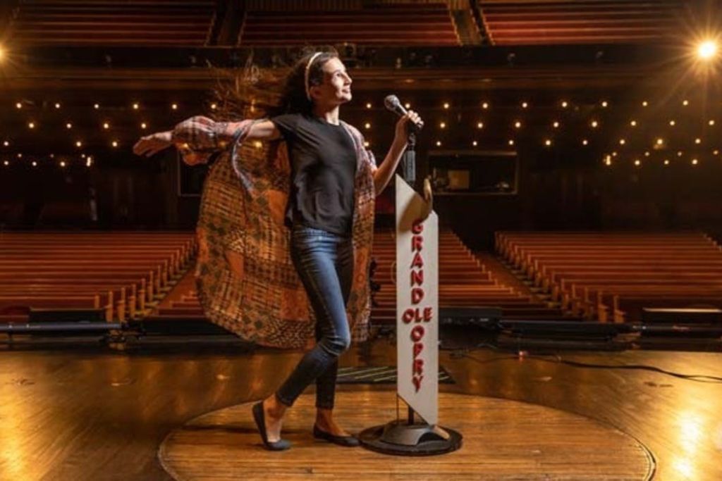 그랜드 올 오프리 (Grand Ole Opry) 무대에서 마이크를 들고 나무 좌석과 조명을 배경으로 극적인 포즈를 취하는 사람.