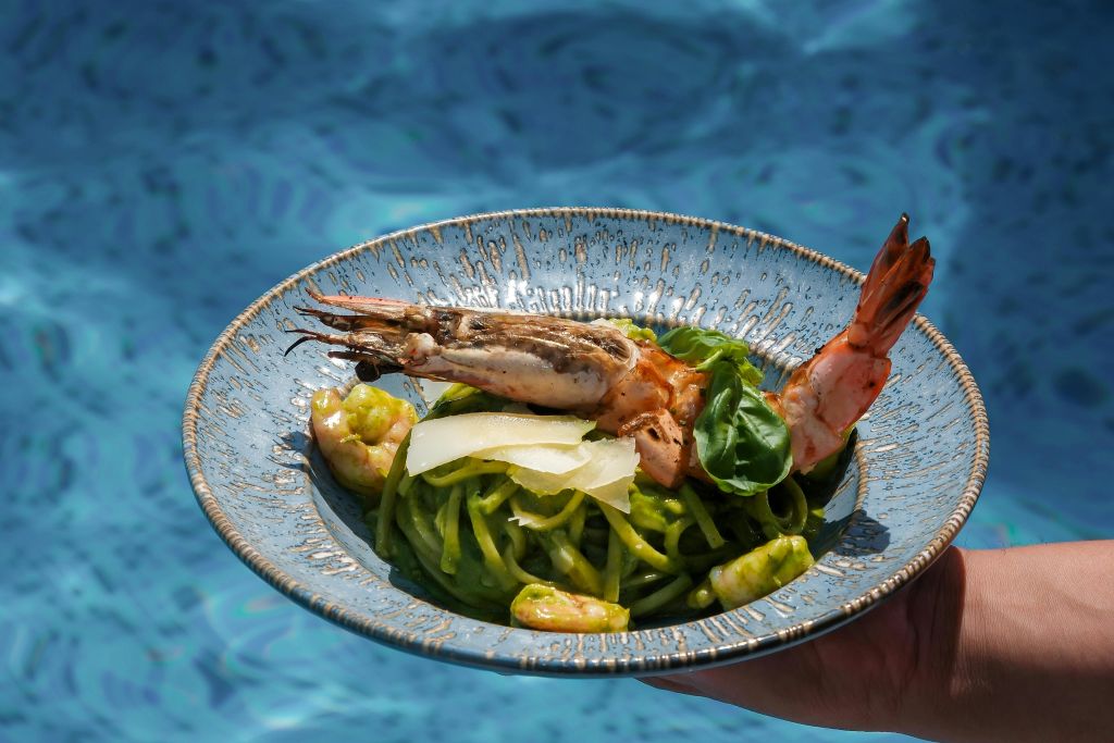 수영장을 배경으로 새우가 담긴 파란색 파스타 접시에 바질과 치즈를 얹은 손이 보입니다.