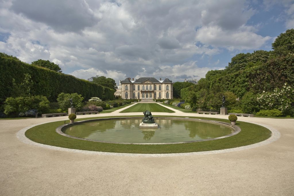 둥근 연못과 조각상이 중심에 있는 정식 정원으로, 구름이 낀 하늘 아래 크고 우아한 저택으로 이어집니다.