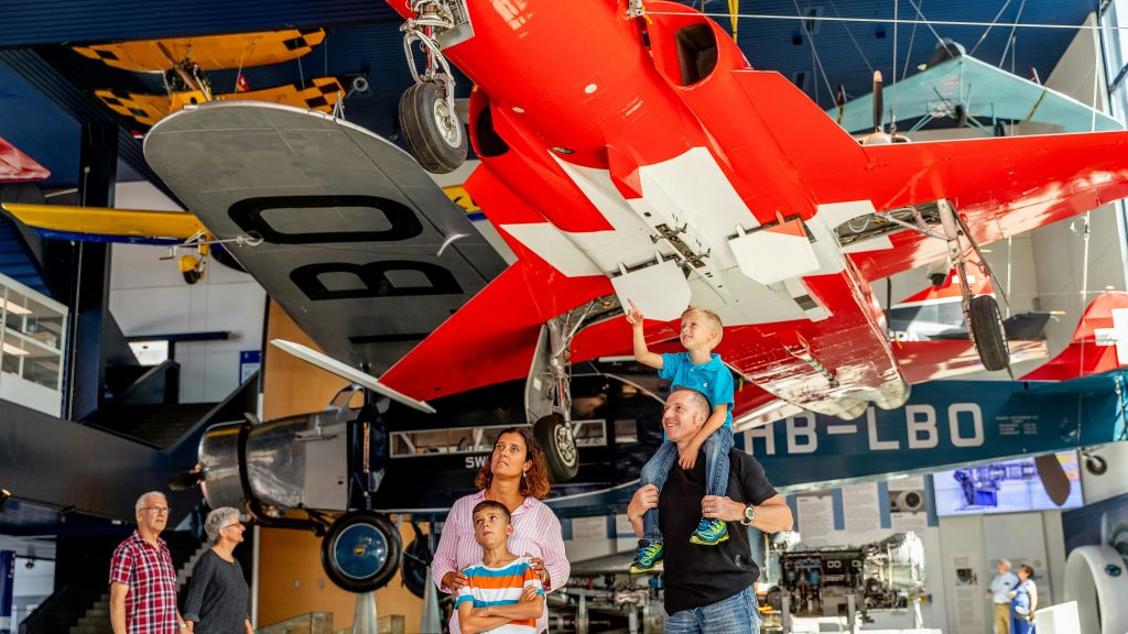 4인 가족이 박물관에 전시된 빨간 비행기를 올려다보고 있으며, 아버지의 어깨에 앉은 아이가 비행기를 가리키고 있습니다.