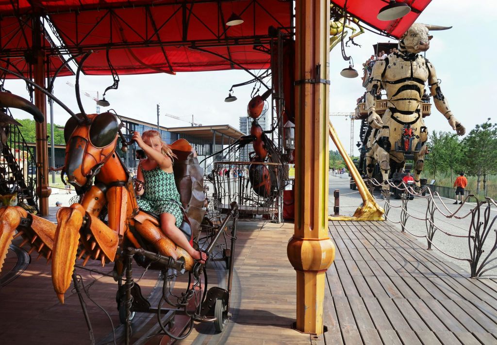 빨간색 캐노피 아래에서 기계 곤충을 타고 있는 아이. 배경에 있는 휴머노이드 피규어의 대형 애니메이션 구조.