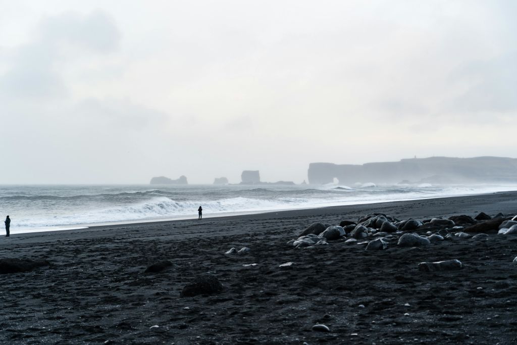 한 고독한 사람이 커다란 바위와 먼 바위 지층이 흐린 하늘 아래 검은 모래 해변에 서 있습니다.