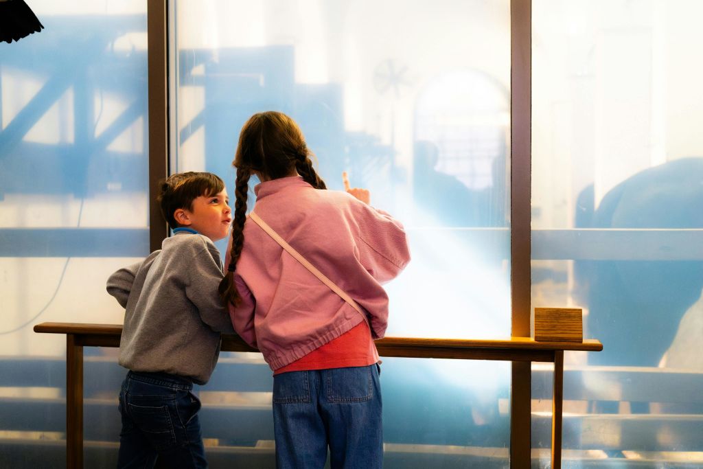 난간 옆에 서 있는 두 명의 어린이가 한 명은 반투명한 창문을 가리키고 있으며, 기계가 배경에 보입니다.