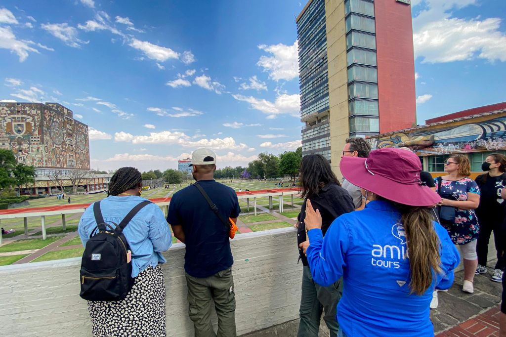 파란색 셔츠와 빨간색 모자를 쓴 투어 가이드를 포함한 일행이 건물을 배경으로 도심 공원이 내려다보이는 벽 옆에 서 있습니다.