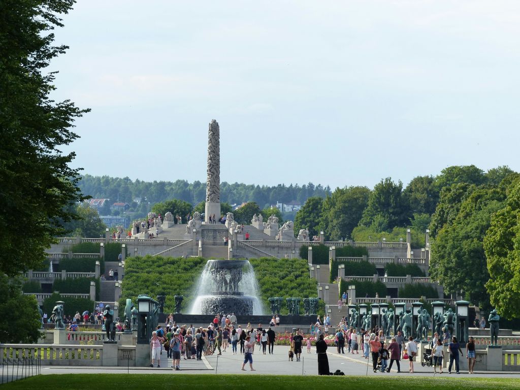 중앙 모노리스가 있는 대형 야외 공원, 전경에 분수가 있고 많은 사람들이 돌아다닙니다. 나무와 녹지가 이 지역을 둘러싸고 있습니다.