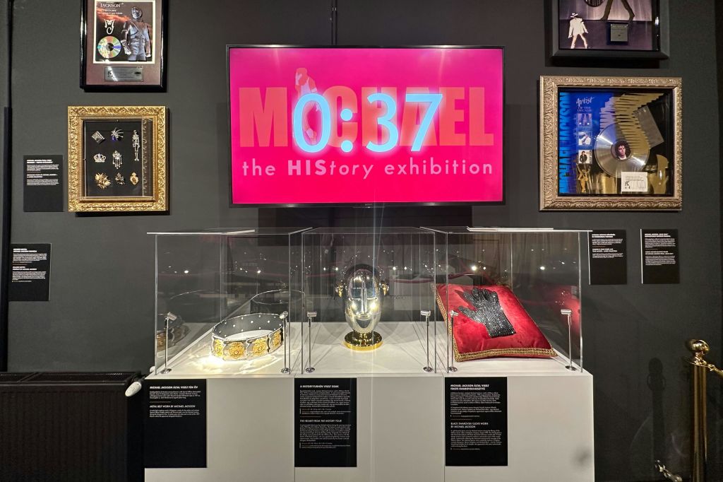 'MJ 0:37 The HIStory Exhibition' 낭독 위에 분홍색 화면이 있는 왕관, 장식용 컵, 유리 케이스의 장갑이 있는 박물관 전시.