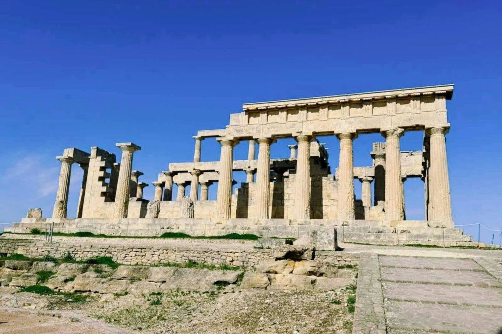 잔디와 고르지 않은 석조 지형으로 둘러싸인 맑고 푸른 하늘 아래 기둥이 있는 고대 석조 사원 유적.