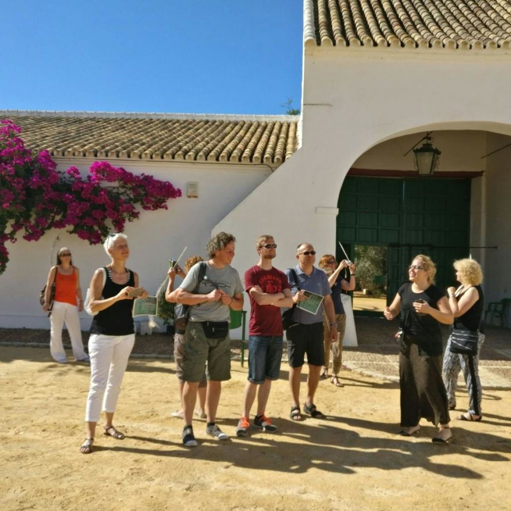 한 무리의 사람들이 아치형 문이 있는 흰색 건물 앞에 서서 라디오를 들고 있습니다. 벽에 화사한 부겐빌레아가 피어납니다.