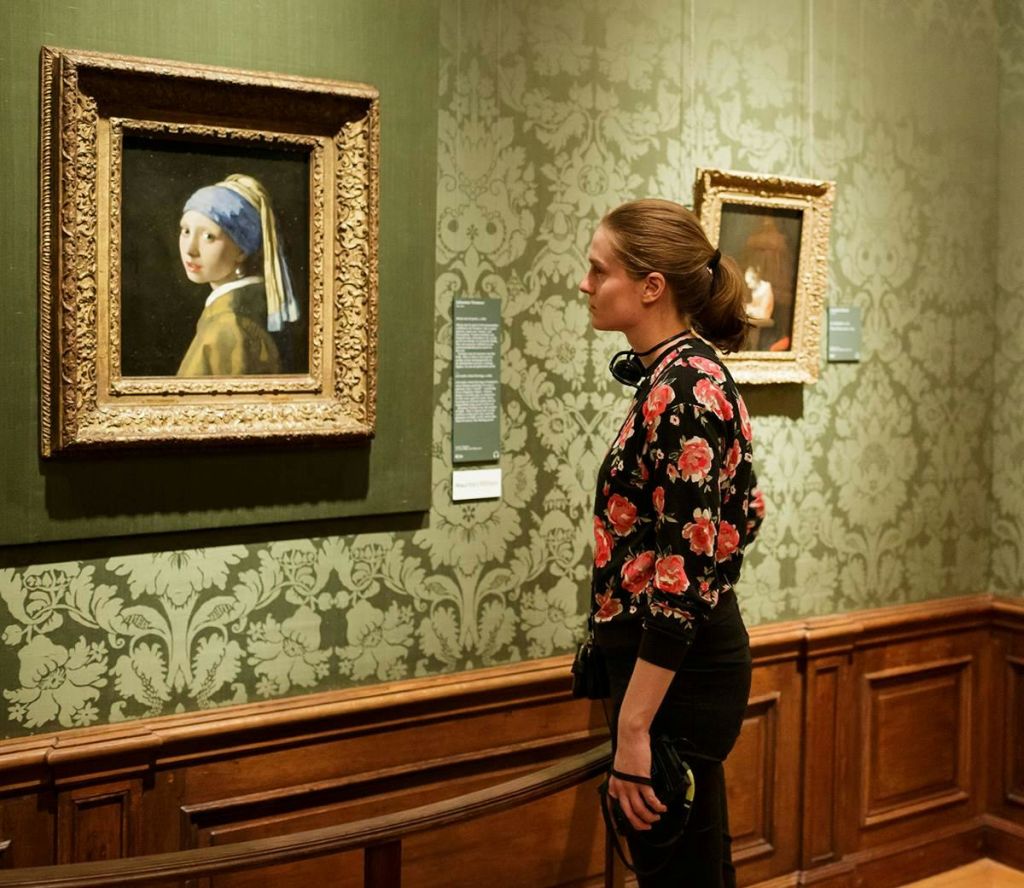 헤드폰을 든 사람이 화려한 벽지와 나무 패널이 있는 박물관 갤러리에 걸린 "진주 귀걸이를 한 소녀" 를 바라보고 있습니다.
