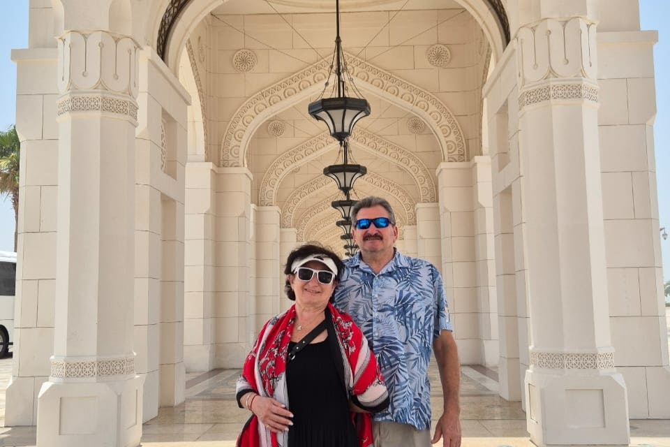 햇살이 내리쬐는 건축 환경에서 선글라스를 쓴 두 사람이 등불이 달린 화려한 아치 아래에서 포즈를 취하고 있습니다.