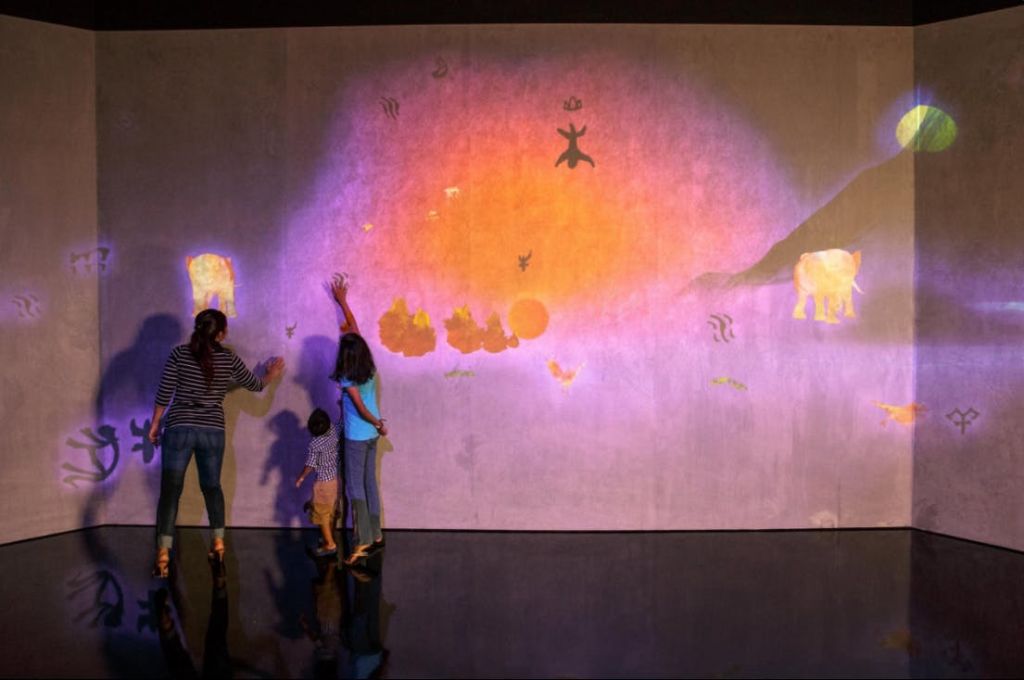 세 사람이 동물 모양과 추상적인 패턴이 특징인 다채롭고 투영된 벽 디스플레이와 상호 작용합니다.