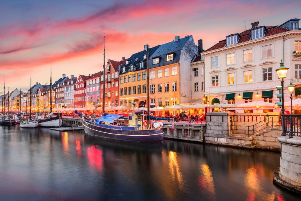 분홍색과 보라색 노을이 지는 하늘 아래 화려한 건물, 정박된 보트, 야외 카페가 있는 물가 풍경.