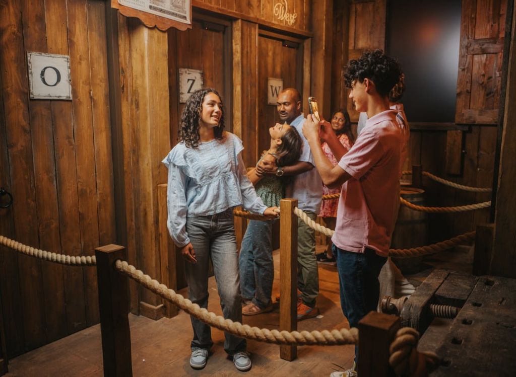 한 사람이 "W" 표지판 옆에서 다른 사람의 사진을 찍고 있는 나무 테마의 탈출실 안의 한 무리의 사람들.