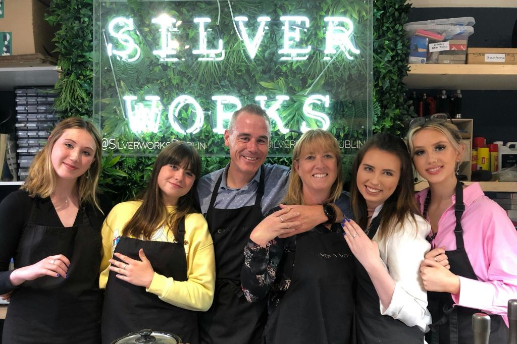 앞치마를 두른 다섯 명이 "Silver Works" 라는 조명 문구와 함께 녹지를 배경으로 미소를 짓고 포옹합니다.