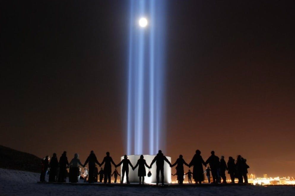 한 무리의 사람들이 비데이 섬의 이매진 피스 타워 주변에서 손을 잡고 있습니다.