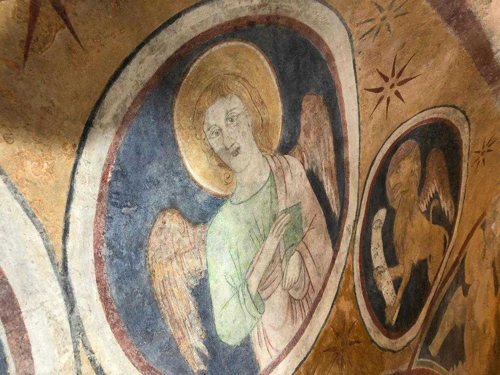 원형 테두리와 별과 같은 장식으로 둘러싸인 후광이 있는 천사가 책을 들고 있는 모습을 묘사한 중세 프레스코화.