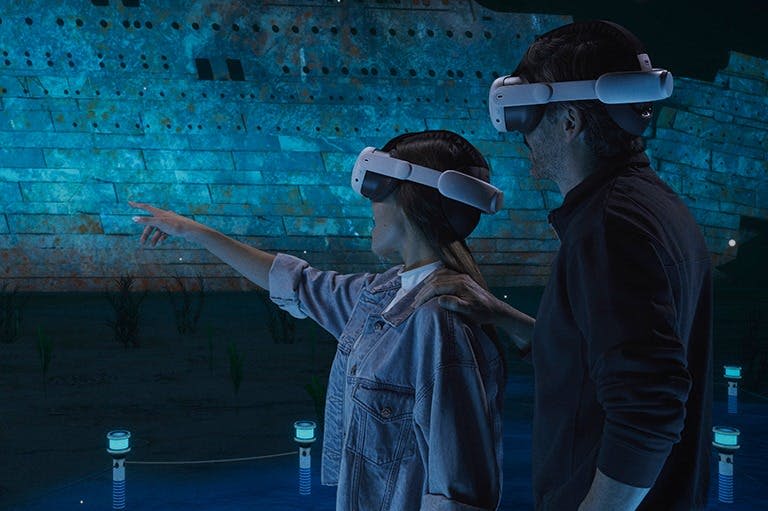 수중 장면과 상호 작용하는 VR 헤드셋을 착용한 두 사람이 커다란 가라앉은 배를 배경으로 등장합니다.