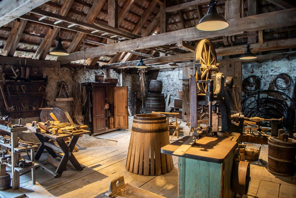 나무 도구, 통, 작업대, 오래된 기계, 램프가 있는 높은 천장 아래 나무 대들보로 가득한 소박한 작업장입니다.