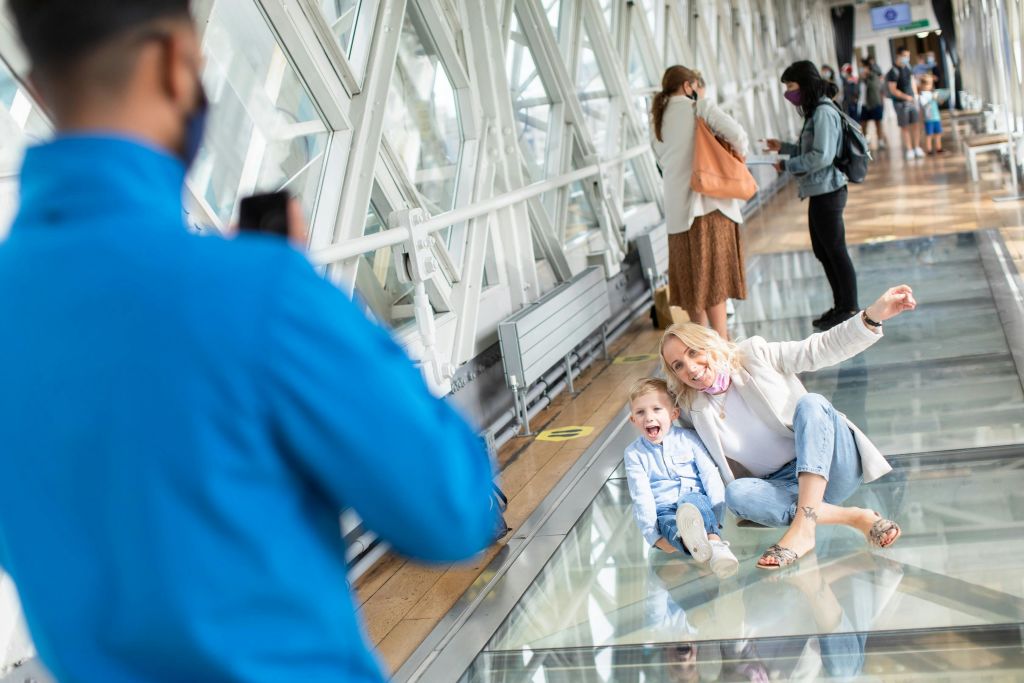 현대적인 실내 산책로에서 한 여성과 어린이가 유리 바닥에서 사진을 찍으며 미소를 짓고 있습니다. 다른 사람들이 걷고 그 지역을 관찰합니다.
