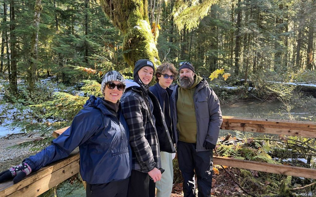 숲이 우거진 지역에서 재킷과 모자를 입은 네 명이 나무 다리에 서서 카메라를 보며 미소를 짓고 있습니다.