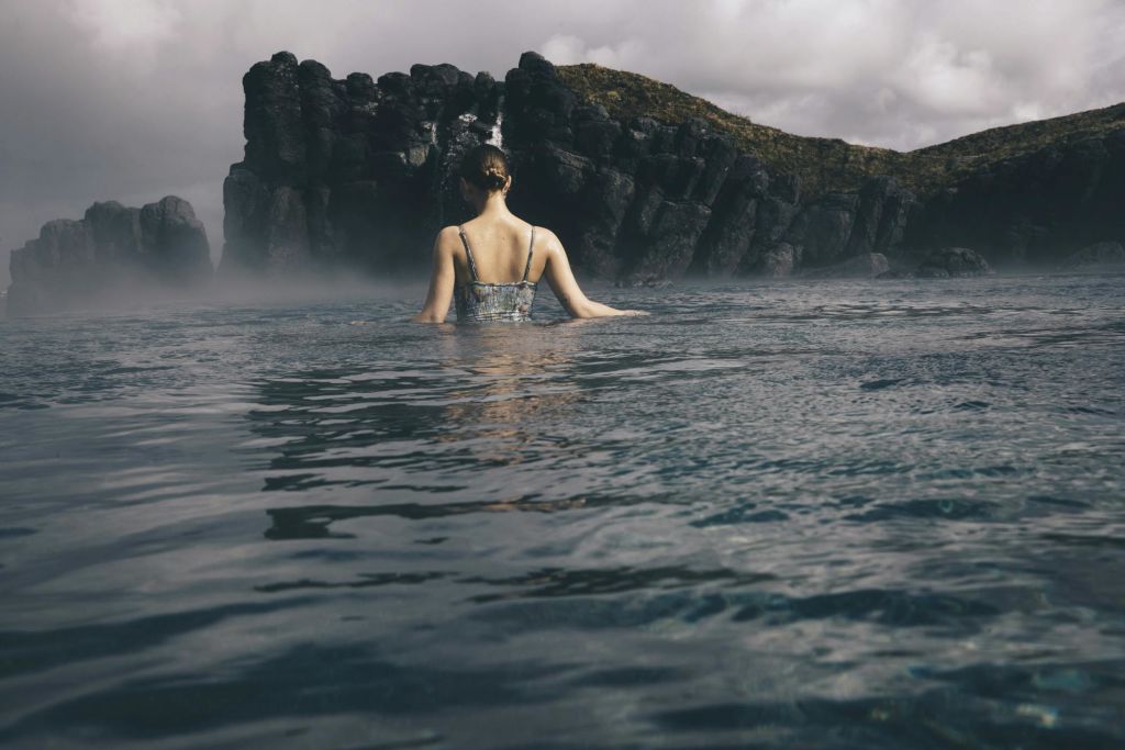 흐린 하늘 아래 커다란 바위와 안개를 배경으로 허리를 물에 담그고 서 있는 사람.