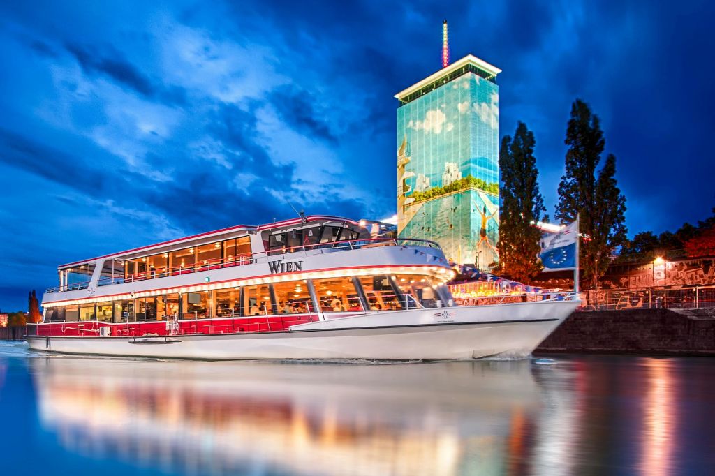 "빈 (Wien)" 이라는 밝은 조명의 강선이 해질녘에 높은 조명 건물을 배경으로 정박해 있습니다.