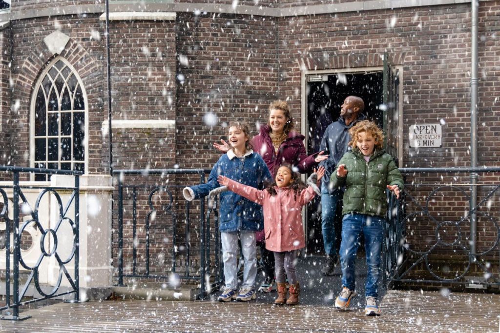 어린이를 포함한 한 무리의 사람들이 벽돌 건물 밖에서 "열기" 라고 적힌 표지판으로 미소를 짓고 적설을 즐기고 있습니다.