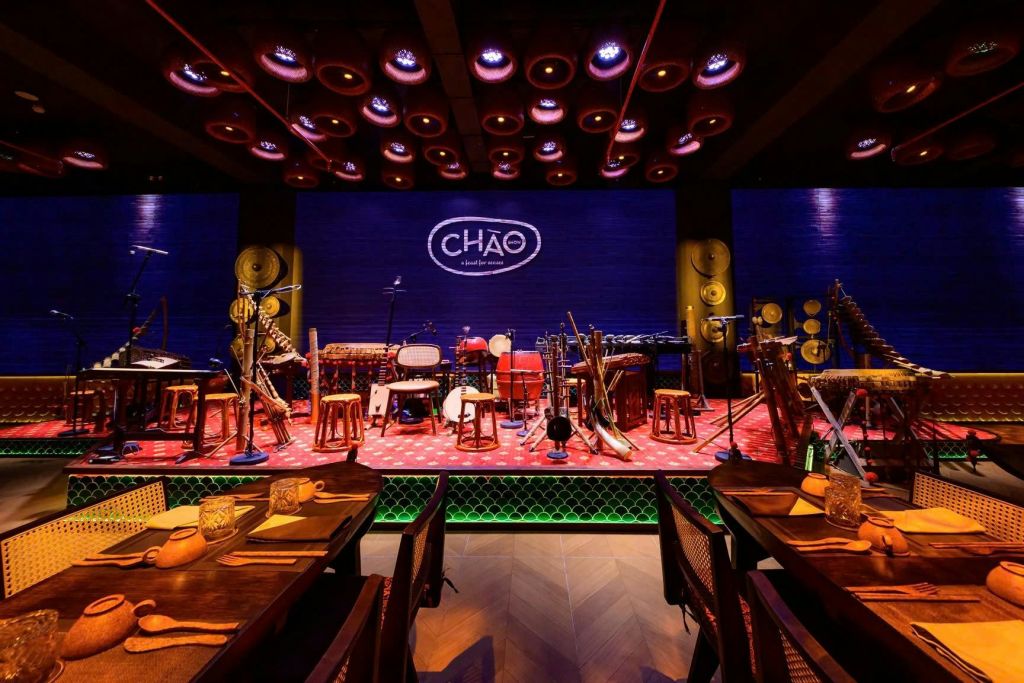 극적인 조명 아래, 탁자, 의자, 파란색 배경에 "CHÁO" 라고 적힌 로고로 둘러싸인 악기가 있는 무대.