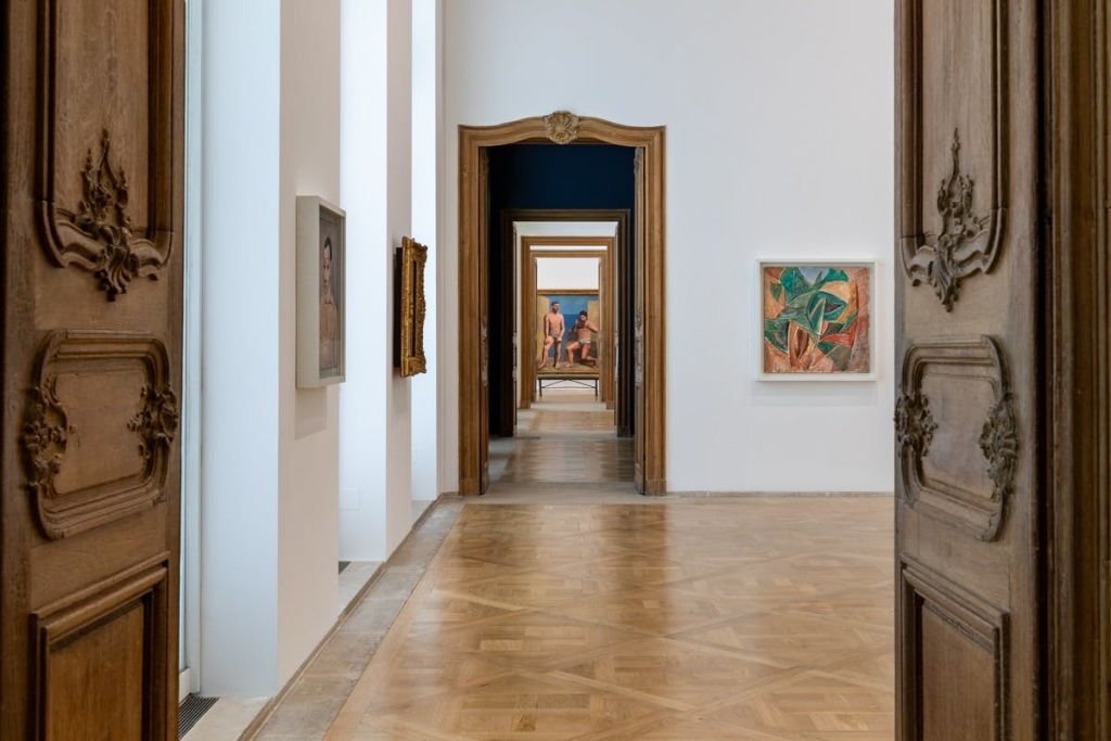 나무 바닥, 흰색 벽, 액자에 담긴 작품, 일련의 열린 출입구가 시각적 깊이를 만들어내는 갤러리 복도.