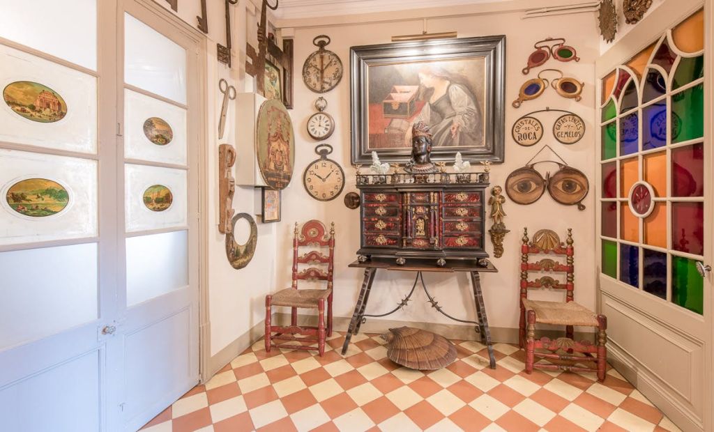체크무늬 바닥, 화려한 의자, 장식된 수납장, 앤티크 시계, 그림 및 기타 빈티지 아이템으로 장식된 벽이 있는 객실입니다.