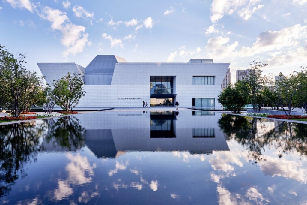 일부 흐린 하늘 아래 나무로 둘러싸인 잔잔한 수영장에 흰색 외관과 커다란 창문이 있는 현대적인 건물.