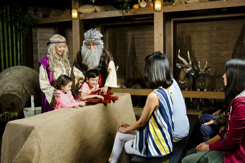 중세 의상을 입은 두 명의 성인과 두 명의 자녀가 나무로 된 역사 도구를 사용하고 다른 여성이 테이블에 앉아 관찰합니다.