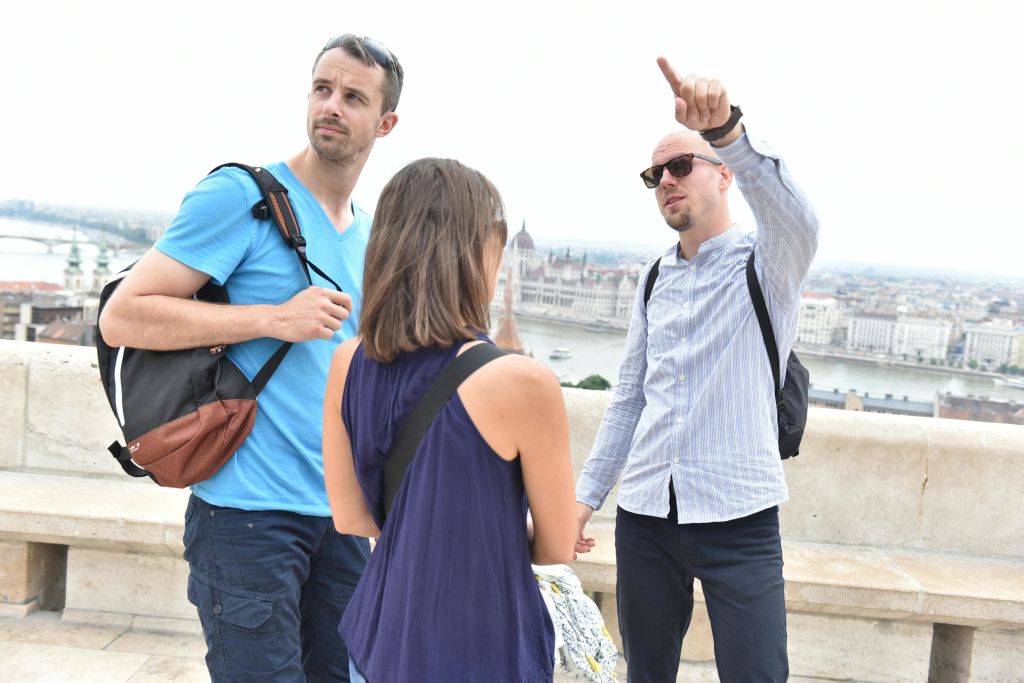 배낭을 든 세 사람이 발코니에 서 있으며, 한 사람이 먼 도시 풍경을 바라보고 있습니다.