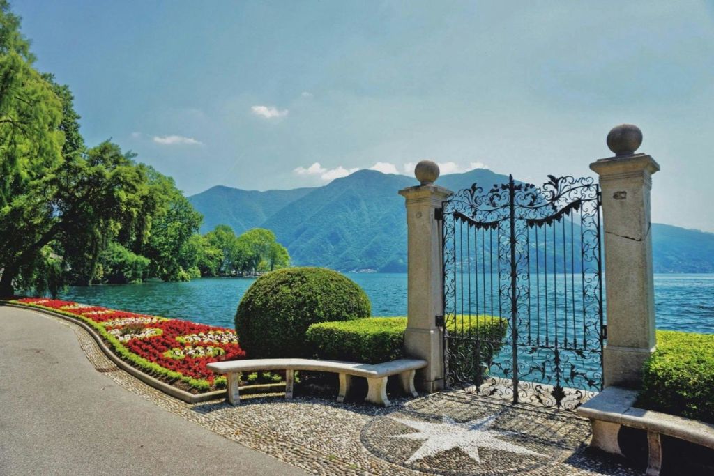산의 풍경을 배경으로 호숫가 정원 옆의 돌기둥이 있는 연철문, 조각된 수풀, 벤치.