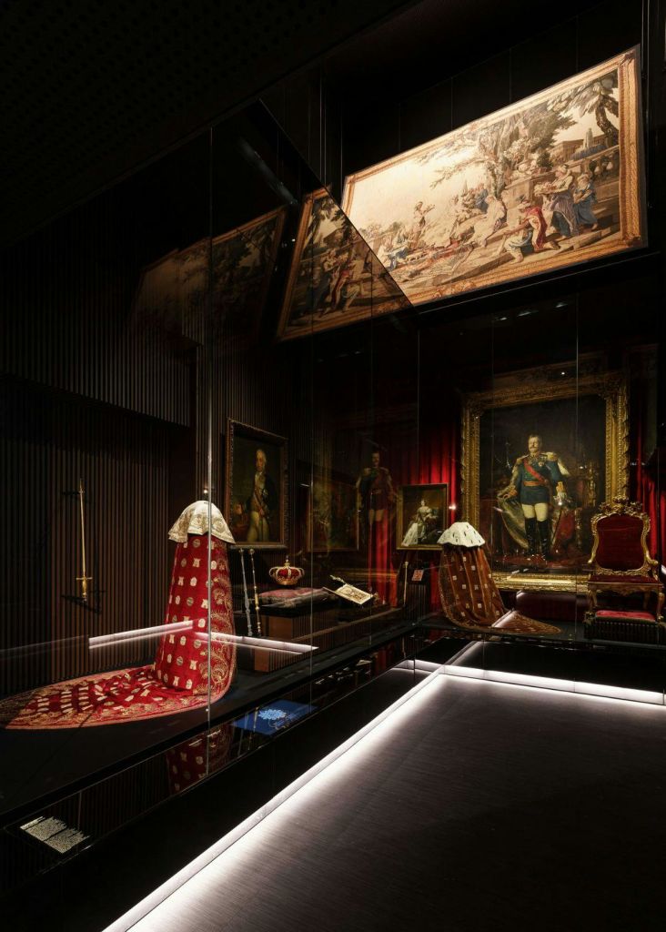 정교한 조명과 어두운 벽으로 장식된 유리 케이스 안에는 왕실 의복, 왕관, 초상화가 전시되어 있습니다.