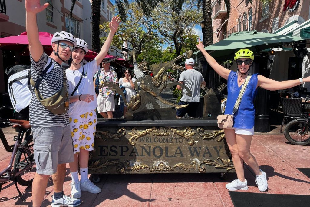 'Welcome to Espanola Way' 표지판이 있는 노란색 헬멧과 파란색 상의를 입은 사람이 사람들과 카페를 배경으로 포즈를 취하고 있습니다.