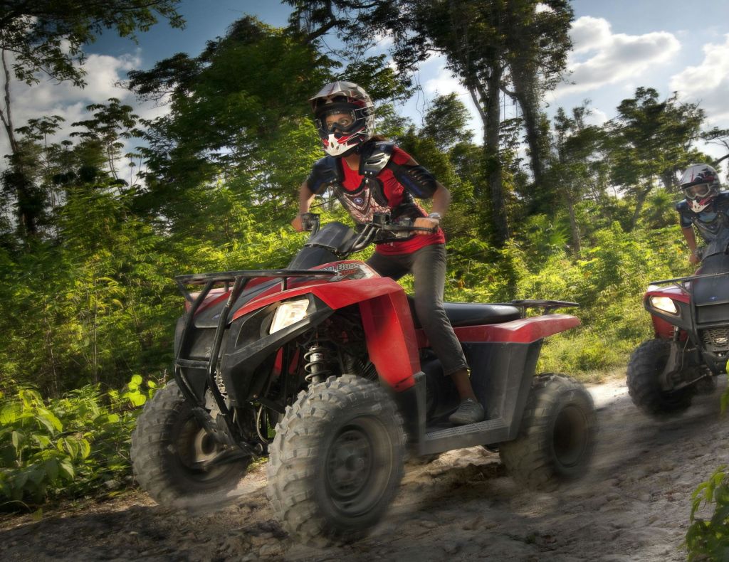 보호 장비를 착용한 사람이 울창하고 푸른 숲을 가로지르는 흙길에서 빨간색 ATV를 타고 있습니다.