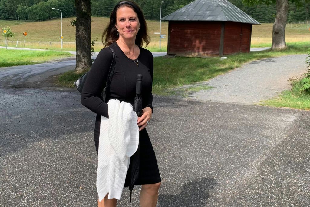 검은색 드레스와 흰색 가디건을 입은 여성이 우산을 들고 잔디와 건물을 배경으로 포장 도로에 서 있습니다.