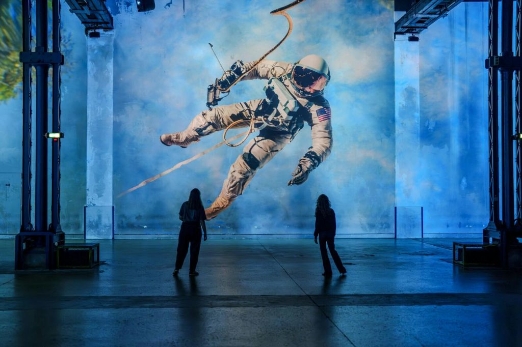 파란 벽에 떠 있는 우주 비행사의 커다란 벽화 앞에 두 사람이 서 있습니다.