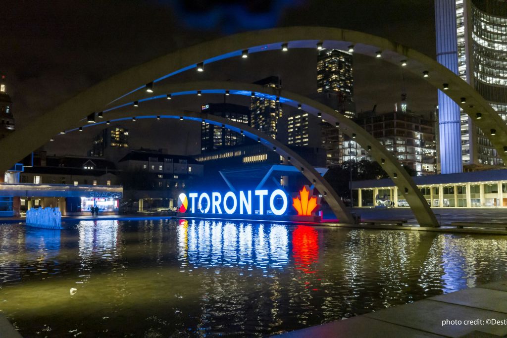 네이선 필립스 스퀘어 (Nathan Philips Square) 의 토론토 표지판은 밤에 불을 밝히고 물 위 콘크리트 아치로 둘러싸여 있습니다.
